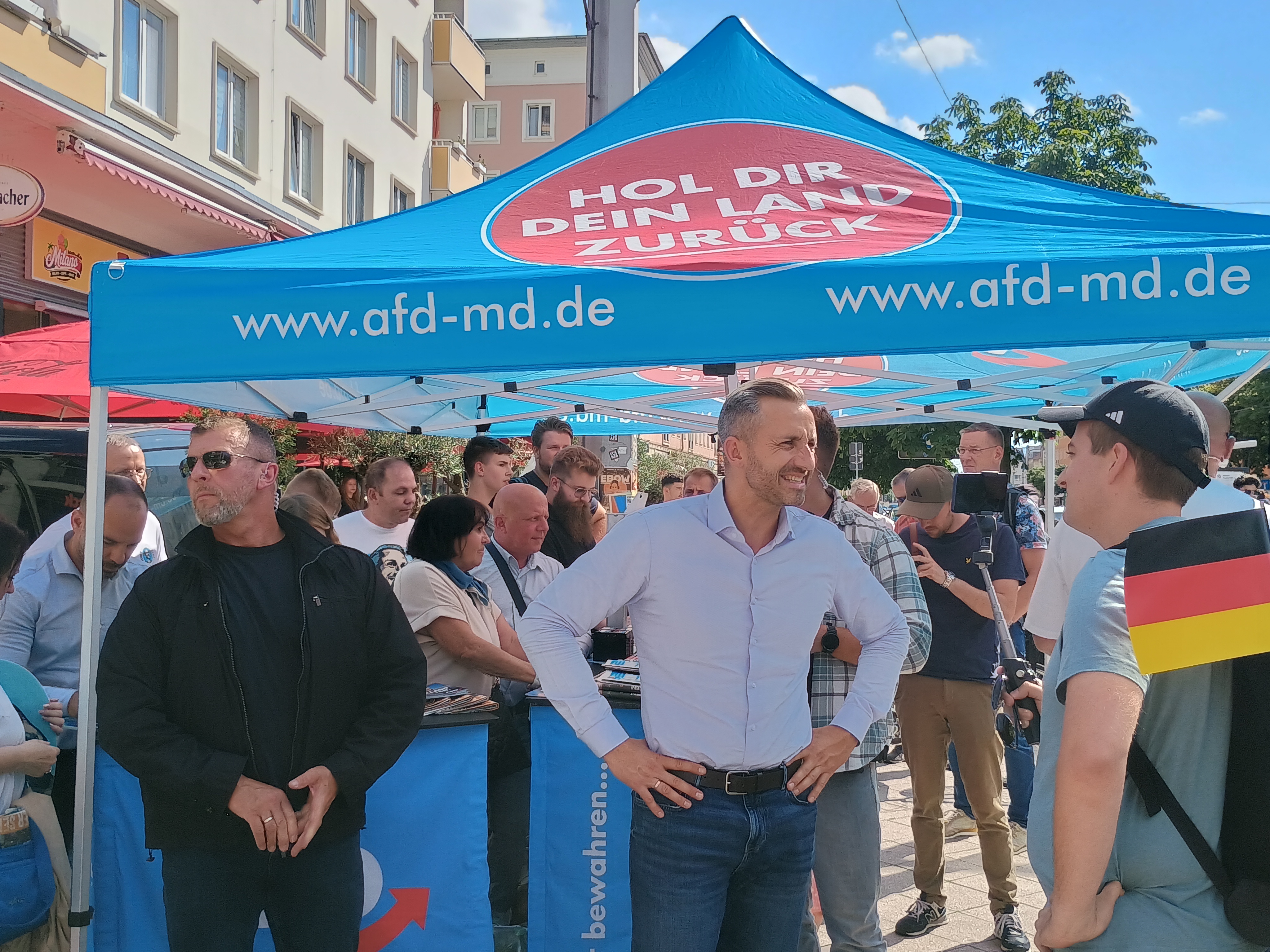 Ulrich Siegmund speaks with supporters beneath an AfD campaign tent bearing the slogan Hol dir dein Land zurück.